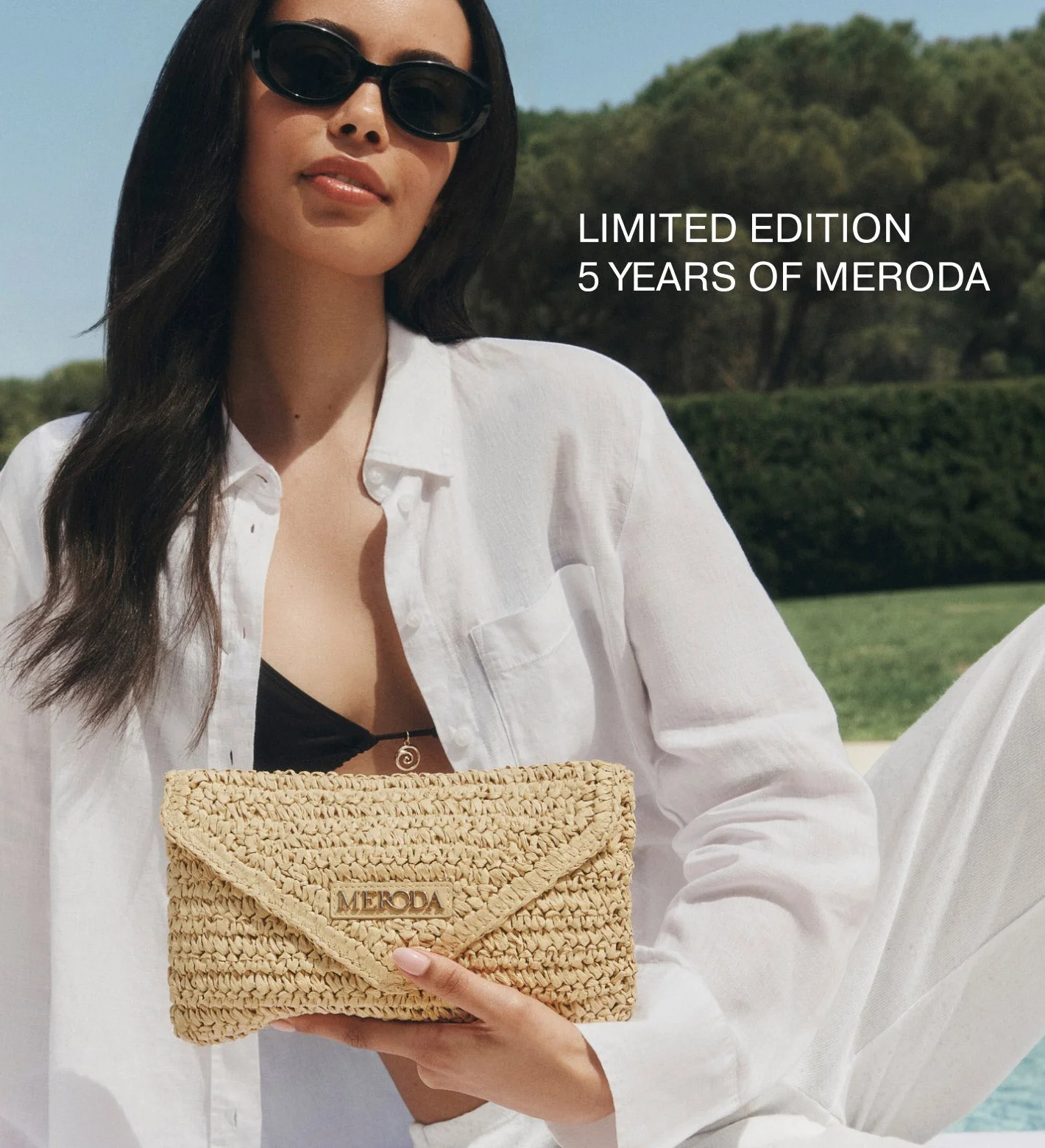 Summer Travel Clutch in woven paper straw held by a model in sunglasses in a sunny outdoor setting.