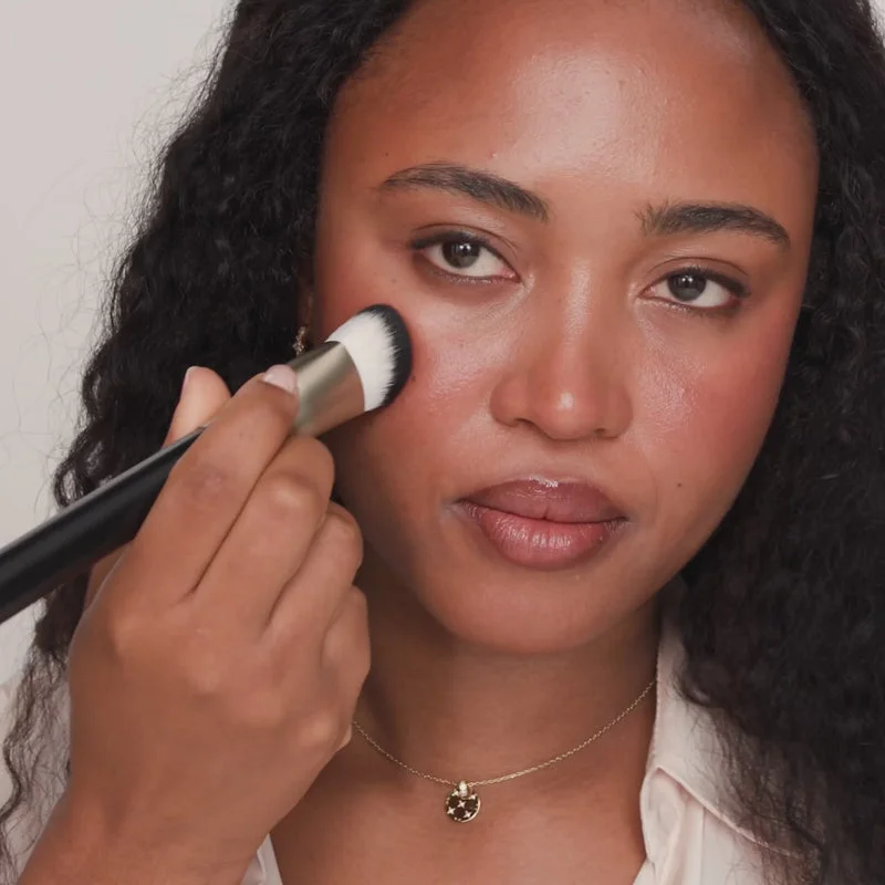 The Essential Brush blending blush on a woman's cheek, highlighting its unique triangular shape and dense bristles.